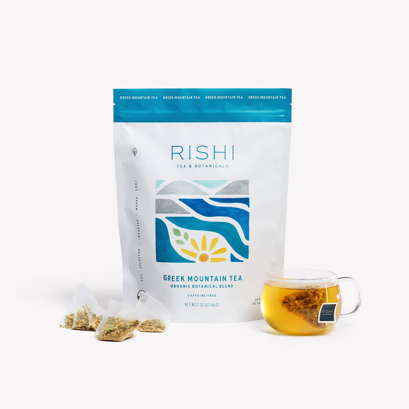 A bag of Rishi Tea & Botanicals Greek Mountain Tea, aromatic ironwort, two tea sachets, and a glass cup filled with brewed tea are displayed against a white background.