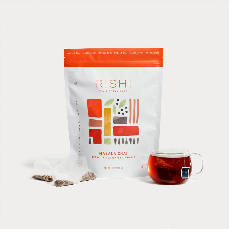 A bag of Rishi Tea & Botanicals Masala Chai organic chai, two tea sachets, and a glass cup of brewed masala chai are arranged on a white background.