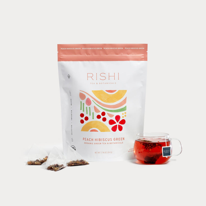 A package of Rishi Tea & Botanicals Peach Hibiscus Green Tea stands behind three pyramid sachets and a glass mug of red tea, showcasing Japanese green tea, hibiscus, and ripe peach flavors on a white background.