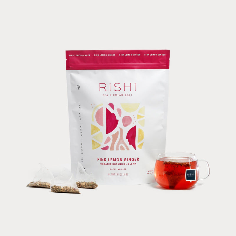 A bag of Rishi Tea & Botanicals Pink Lemon Ginger tea—a vibrant botanical blend with shiso—stands behind three sachets and a clear glass cup of brewed tea.