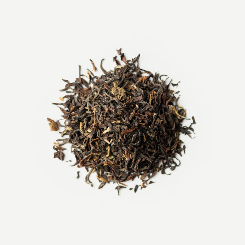 A small pile of Darjeeling Second Flush Chamong DJ73 by Rishi Tea & Botanicals on a white background. The dried loose-leaf tea showcases green and brown hues, with subtle hints of stone fruits interwoven among the fragrant blossoms.