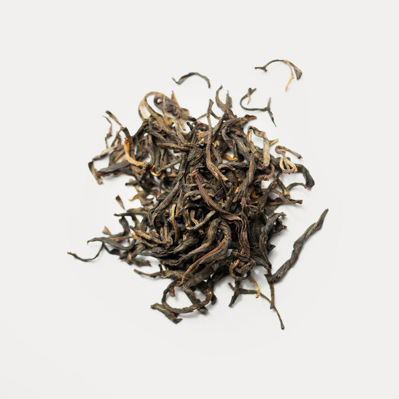 A pile of Rishi Tea & Botanicals loose leaf Dian Hong black tea with twisted, dark brown and golden leaves promises a rich, malty flavor against a plain white background.